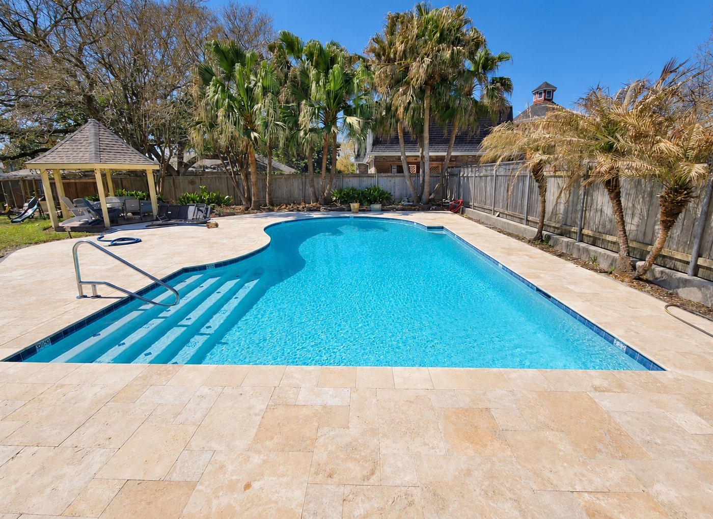 Pool with clear blue water, tiled steps, a handrail, and a travertine deck and coping, with a gazebo and palms behind it