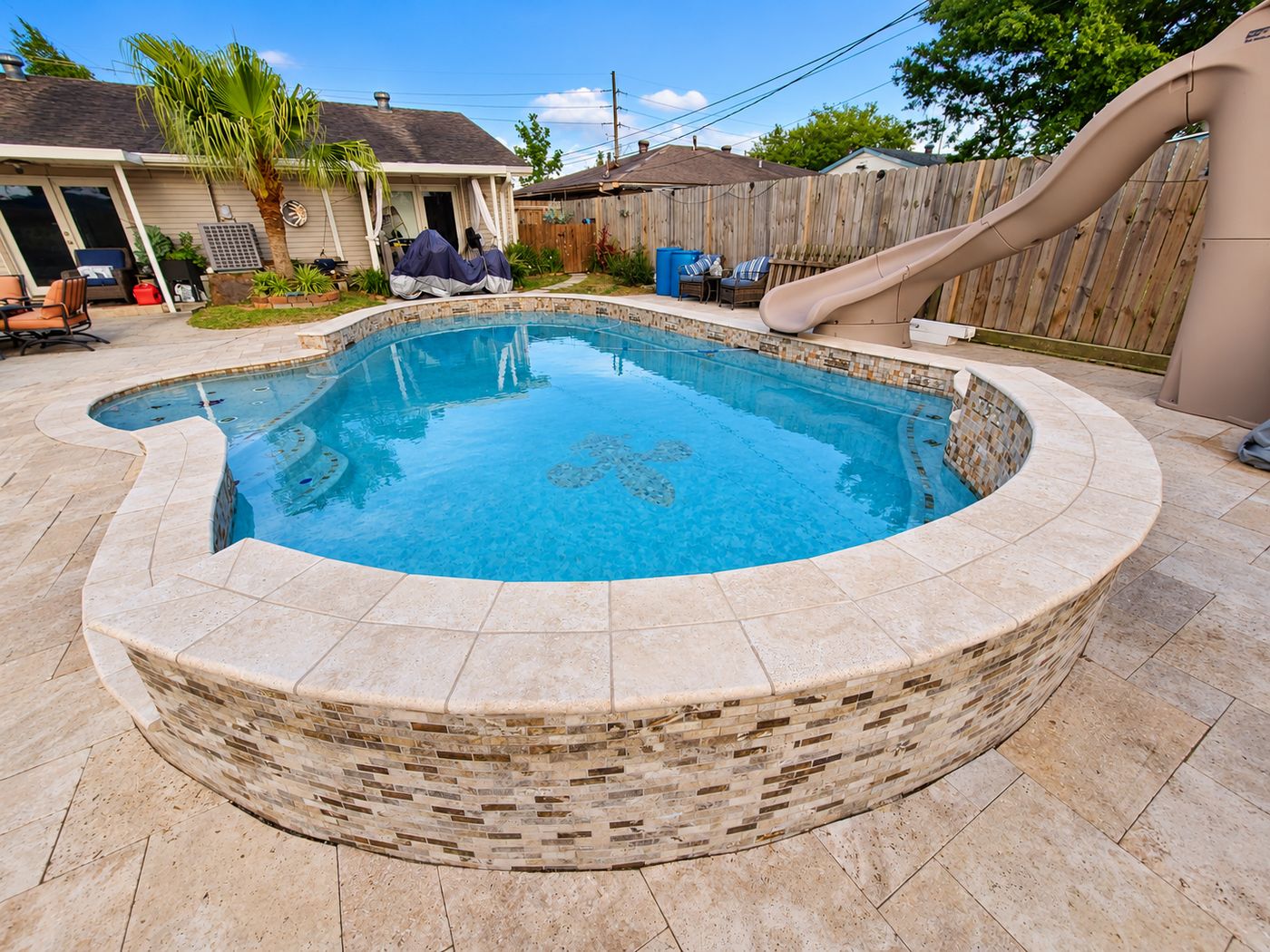 Raised freeform pool with a fleur-de-lis mosaic on the floor, a tile-faced wall, travertine coping and deck, and a water slide
