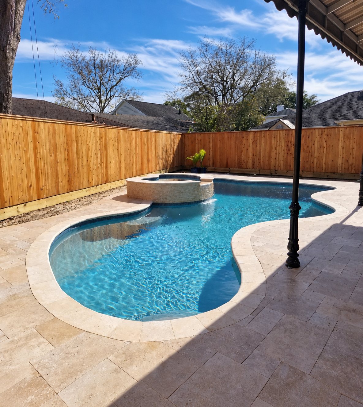 Freeform pool with a raised stacked-stone spa spilling into it, surrounded by a travertine deck and a new wood fence