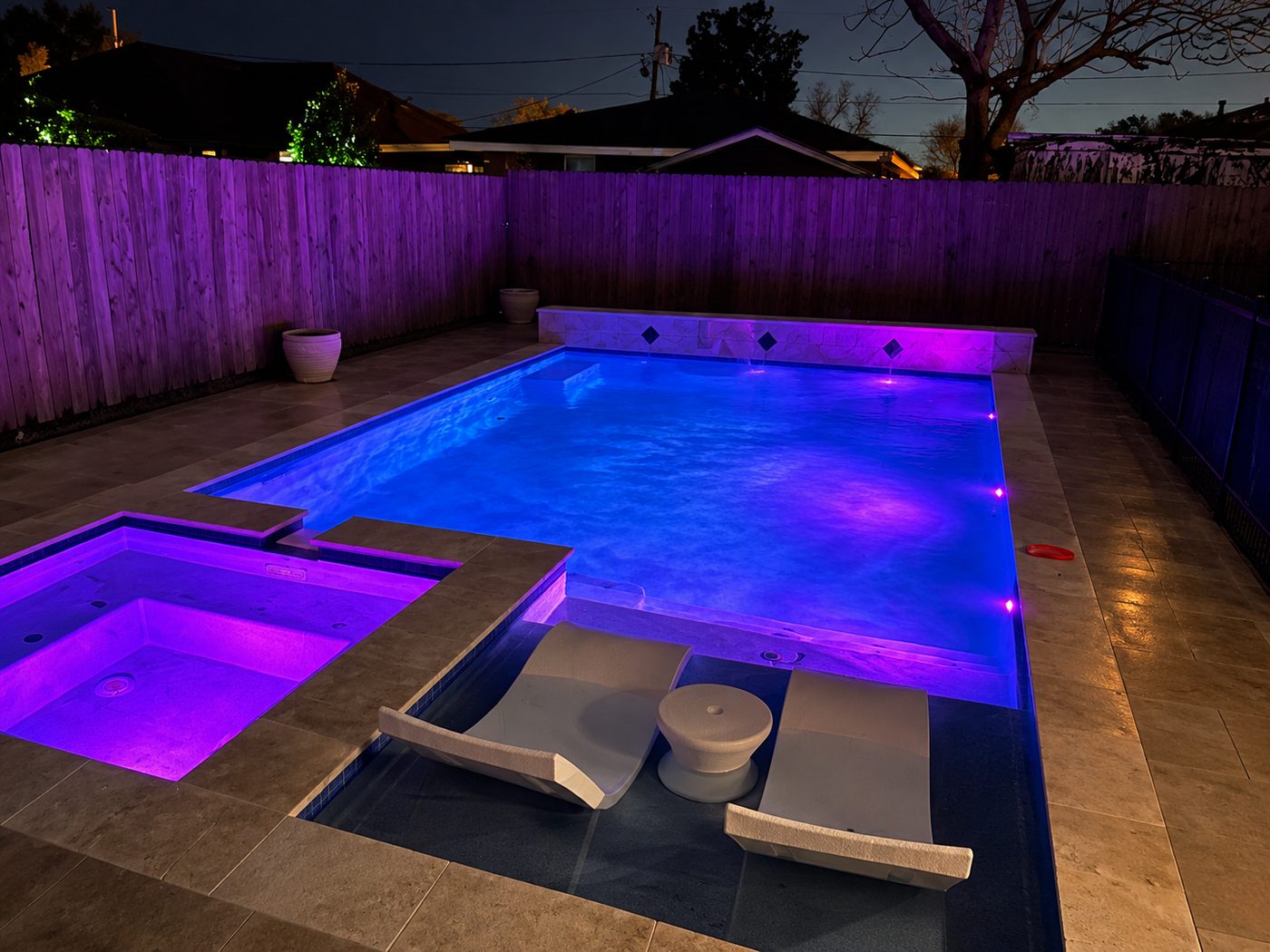 Pool and spa lit purple and blue at night, with in-pool loungers on the sun shelf and a stone water-feature wall